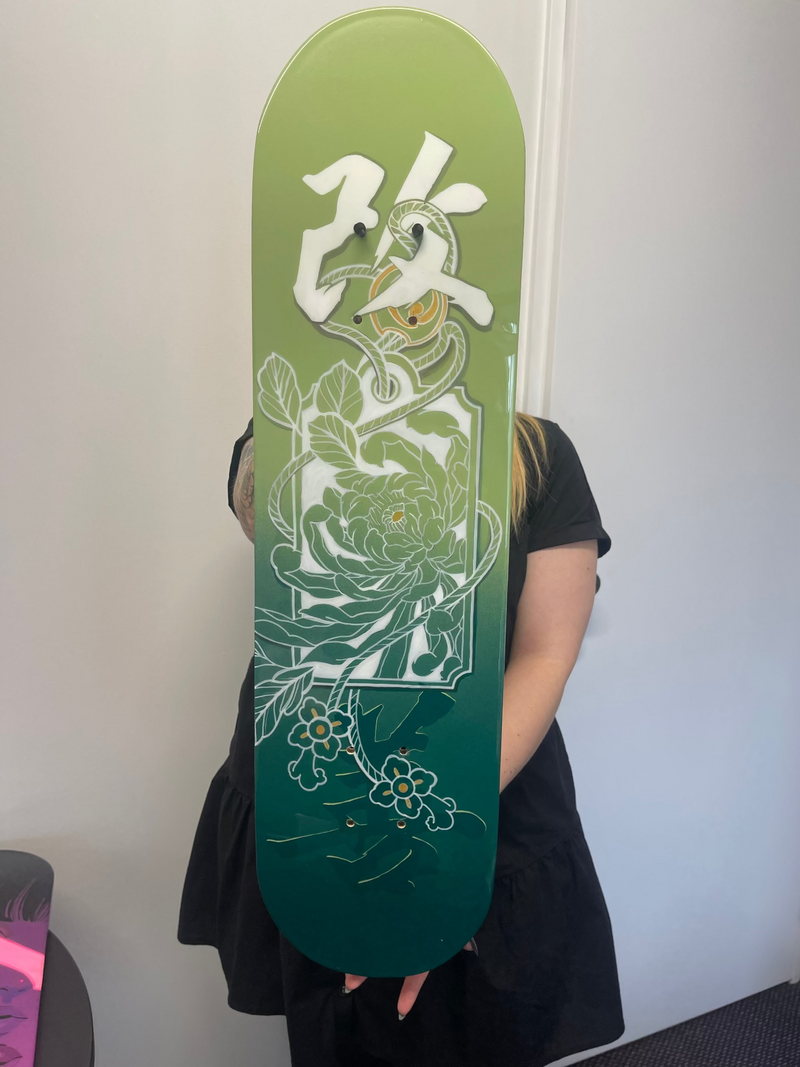 A person in a black dress holds a skateboard deck featuring a green gradient design with white floral illustrations and a large white character graphic.