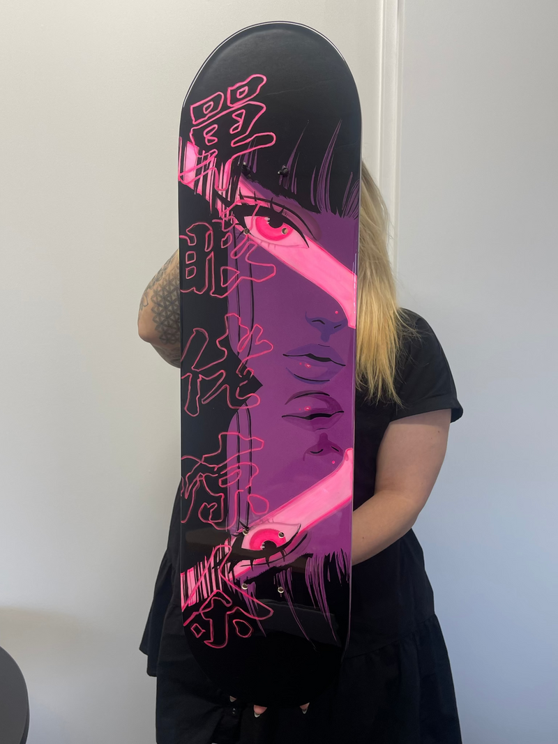 A person holding a black skateboard with a bold graphic design featuring a stylized face in purple and pink, with pink Japanese characters, against a plain white wall.