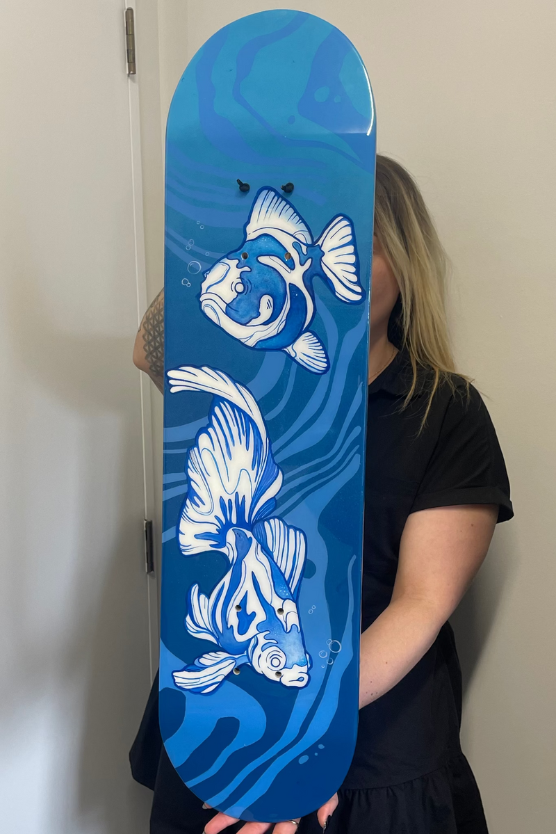 A person holding a skateboard deck featuring a blue water design with two white koi fish, photographed indoors against a plain wall.